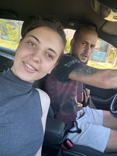 A rotated, bright daylight selfie captures two individuals inside a car, likely in Rogozen, Bulgaria. In the foreground, a woman with dark hair pulled back smiles broadly at the camera. She has light brown eyes, a fair complexion with freckles, and wears a dark grey, ribbed, high-neck top, conveying a cheerful and relaxed mood.
Behind her, a man with a bald head or very short hair and a beard looks towards the camera with a subtle, neutral smile. He is wearing a dark t-shirt with an abstract, patterned design, and a prominent tattoo is visible on his right arm. His lower half shows white shorts that appear somewhat soiled.
The car's interior features dark seats, and a red seatbelt buckle with the word "PRESS" is clearly visible next to the man's leg. Through the windows, a sunny outdoor environment is seen, including bright green foliage, a metal fence, and what appears to be a building or gate, suggesting a rural or suburban setting. A partial word, possibly "WASH," is visible on the man's t-shirt.
