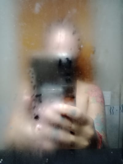 A blurry image shows someone in a bathtub. A tattooed arm rests near the edge of the tub, with a hand partially submerged. There's an orange tint to the water, possibly from a bath product. Steam or condensation obscures much of the scene, but a black rectangular shape, likely a phone or device, is visible in the water.