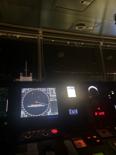 The image shows the interior of a ship's bridge at night. Various screens display nautical information like radar and navigation data. An external view of city lights along the water is visible through the window. Control panels with gauges and buttons are prominent features, highlighting the technological aspect of maritime navigation. The environment is dark, with light primarily emanating from the screens and control panel displays.