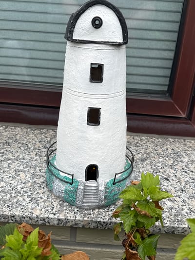 A ceramic lighthouse figurine stands on a speckled gray windowsill in front of a closed gray-slatted window and a dark red window frame. The lighthouse is white with a black top and a black rim around the domed roof. It has two square window openings on its tower and a small arched doorway at the base with stairs leading up to it. A black metal railing encircles the base of the lighthouse. To the right of the lighthouse, a small green plant with some brown, dried leaves is visible. The lighthouse is positioned in Bassum, Germany.