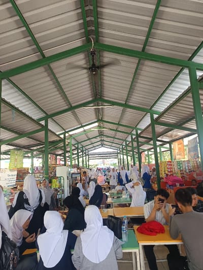 An open-air cafeteria-style space is filled with people, predominantly women wearing white hijabs. They sit at tables, some eating, others talking or looking at phones. The structure has a metal roof supported by green metal beams. One or more ceiling fans are visible. A snack shop lines part of the area, displaying goods for sale. Natural light floods the space.