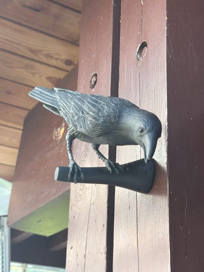 A dark gray, ornamental bird figurine, possibly a crow or raven, is mounted on a dark brown wooden post. The bird is positioned facing towards the right of the frame, with its beak lowered as if pecking at something. Its wings are detailed with a feathered texture. The bird is perched on a dark, cylindrical object attached to the post. The post itself has visible wood grain and appears to be part of an outdoor structure, possibly a porch or fence. In the background, there is a lighter brown wooden ceiling or overhang. The image appears to be taken outdoors, with natural light illuminating the scene. The overall setting does not provide any specific indicators for location within Nizhnyaya Shilovka, Russia.