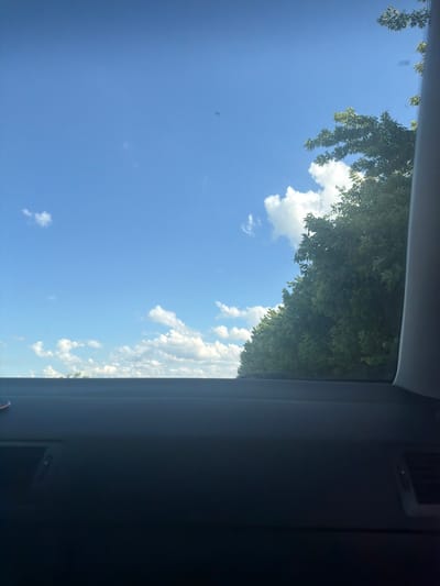 The image captures a view of the sky from inside a vehicle, likely on a sunny day. The perspective is from the passenger seat, looking upwards through the windshield and side window.

The primary subjects are the bright blue sky dotted with fluffy white clouds, and the lush green foliage of trees that overhang the view. The trees are in full leaf, suggesting a warm season, likely summer or late spring. Some branches and leaves are visible in the upper left and upper right portions of the image, creating a natural frame.

The scene suggests a journey or a moment of pause during travel. The setting appears to be outdoors, with the sky and trees indicating a location with natural surroundings, possibly a rural or suburban area. There are no people visible in the image, and the only objects are the natural elements and the interior of the car, which is partially visible as a dark, shadowed area on the right side of the frame. A rectangular object, possibly a dashboard element or a vent, is subtly visible in the upper right corner within the car's interior.

The weather is clearly sunny with good visibility, given the brightness of the sky and the distinct white of the clouds. The time of day is likely daytime, given the blue sky.

There is no visible text in the image. The overall emotion conveyed is one of peace and natural beauty, perhaps a moment of enjoying the scenery during a drive. The crops provided offer closer views of the sky, clouds, and foliage, reinforcing these observations.