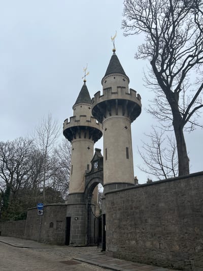 The image shows the towers of St. Machar's Cathedral in Aberdeen, Scotland, United Kingdom. The two prominent towers are made of light-colored stone with decorative features and conical roofs topped with weather vanes. Bare tree branches frame the towers against a cloudy sky. Part of a stone wall extends from one of the towers and runs alongside a paved road. A signpost with a blue circular sign displaying bicycle symbol and a rectangular sign with an image of a camera above a coin slot is also visible near the base of the tower.