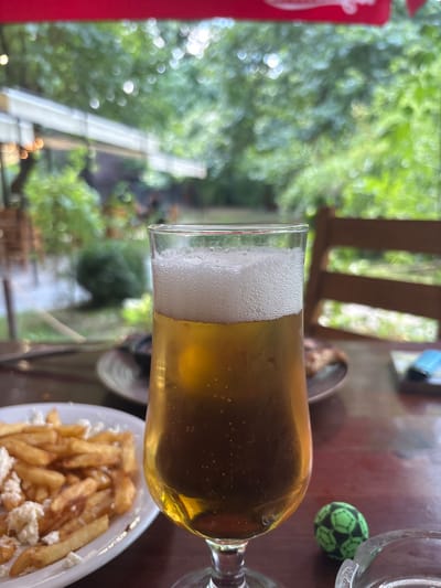 The image is a close-up shot of a glass of beer on a wooden table, with a plate of french fries in the foreground to the left. In the background, there is a blurred outdoor setting with trees and greenery, suggesting a restaurant or beer garden. The overall ambiance is casual and relaxed, likely taken at an outdoor dining establishment in Parvenets, Bulgaria, possibly during daylight hours given the natural light illuminating the scene. A small green and black soccer ball is visible on the table near the beer glass.