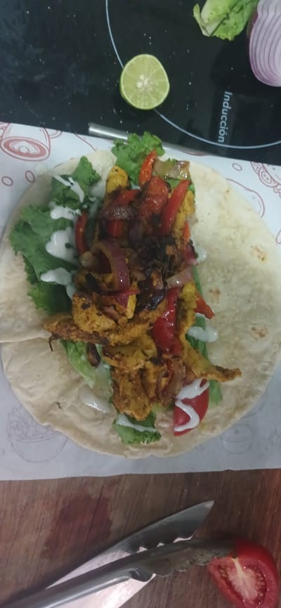 A wrap filled with chicken, peppers, onions, lettuce, and sauce sits on a white paper liner, which is on a black stovetop with a lime slice and half an onion nearby. Below the stovetop, a wooden cutting board holds a pair of tongs gripping a tomato slice. The scene is likely in Veracruz City, Mexico, given the context.