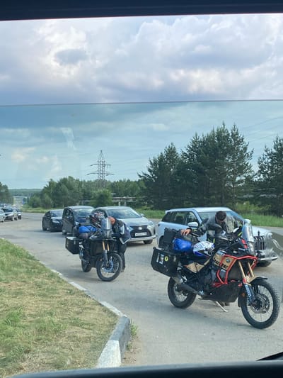 The image shows a traffic jam on a road, likely in the city of Belorechensky, Russia, as indicated by the license plate format on one of the vehicles. The sky is overcast with scattered clouds, and the road is lined with trees and grass. Several cars are stopped, and two adventure motorcycles are prominently featured in the foreground. One motorcycle is parked on the right side of the road, with a person in riding gear leaning over it. This motorcycle has saddlebags and a helmet mounted on it. Another similar motorcycle is positioned behind it, also with luggage. The traffic appears to be backed up on a two-lane road. A power line pylon is visible in the background, suggesting the presence of infrastructure. The overall scene depicts a delay in traffic flow, with motorcyclists possibly taking a break or assessing the situation.