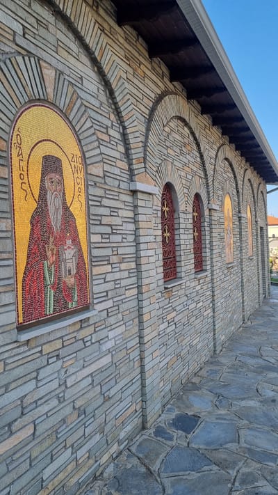 The image shows a close-up, angled view of the exterior of a building with a stone facade. The building features several arched openings, some containing decorative red metal grilles with cross motifs, and others housing mosaics. The most prominent mosaic on the left depicts a robed figure with a long beard, holding a building-like object, against a golden background. The stonework is layered and varied in color, ranging from grey to tan. A tiled walkway is visible in the foreground, leading away from the building. The scene is illuminated by natural light, casting subtle shadows. The overall impression is of an outdoor religious or historical structure in a Mediterranean setting, likely Litochoro, Greece, given the architectural style and mosaic elements common in Orthodox Christian churches.
