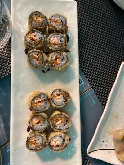 This close-up shot captures a white rectangular plate filled with twelve deep-fried sushi rolls, arranged in two rows of six. Each roll is golden-brown from frying, suggesting a crispy exterior, and has a dark, glossy sauce drizzled over it. The visible cross-sections of the rolls reveal white rice, dark seaweed (nori), and a filling that appears to include pinkish-orange fish (likely salmon) and a lighter, creamy ingredient, possibly cream cheese.

The plate rests on a dark gray placemat with a repeating textured pattern. In the upper right corner, a portion of another white ceramic dish with a textured surface and some light-colored food (possibly dumplings or a similar appetizer) is visible. A clear glass is partially seen in the bottom left corner.

The scene depicts a dining experience, likely at a restaurant or eatery in Basiglio, Italy. The lighting suggests an indoor setting, possibly evening, with artificial light illuminating the food. There are no people visible, and no discernible text appears in the image.