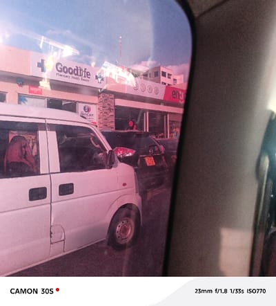 The photo, shot from inside a car, features a white van and a dark vehicle with a yellow license plate. A building with a "Goodlife Pharmacy Health Beauty" sign is visible. Reflections and a pinkish hue affect the image quality. The photograph was taken with a CAMON 30S camera at 23mm f/1.8, 1/33s, ISO770.