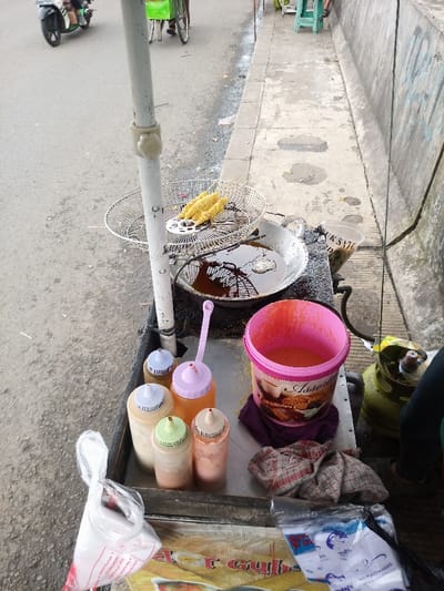 This image presents a street food vendor's stall positioned at the edge of a paved road and an adjacent concrete sidewalk in Caringin, Indonesia. The stall features a dark, elevated surface, serving as a cooking and serving area. A wok-style pan filled with dark cooking oil is visible, reflecting ambient light. Next to the pan, a wire mesh frying basket holds several golden-brown, elongated fried items impaled on wooden skewers, suggesting they are either freshly cooked or draining.
Six plastic squeeze bottles with varied colored caps (blue, purple, orange, light green, and beige) are arranged on the surface, containing different sauces or condiments. A pink plastic bucket, labeled "Assorted," contains a pinkish-orange liquid, likely batter, with a pink ladle resting within it. A crumpled red and white plaid cloth and a plain purple cloth are also on the surface. On the lower right, a green LPG gas cylinder is partially visible, supplying fuel for the cooking operation. The lower legs and feet of an individual, wearing dark pants and sandals, are seen standing next to the stall.
In the background, on the paved road, a person rides a black motorcycle, and further away, another person is seen operating a green bicycle cart. The concrete sidewalk runs parallel to the road, bordered by a concrete wall displaying faint blue graffiti. The overall scene indicates active street food preparation and vending during daylight hours.