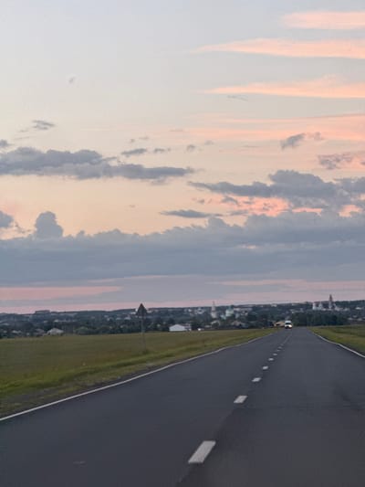 The image depicts an asphalt road with white lane markings, stretching into the distance. The road is bordered by a grassy field on the left and a line of trees and buildings on the right, suggesting a semi-rural or suburban setting. In the background, a town or village can be seen nestled among the trees. The sky above is filled with a mix of light and dark clouds, with hints of pink and orange, indicating either sunrise or sunset. The overall atmosphere is peaceful and serene. There are no people visible in the image. The time of day is likely either early morning or late evening. No text is visible.