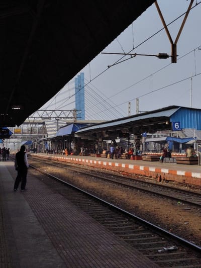 The image displays an outdoor scene at Bardhaman Junction railway station in Bardhaman, West Bengal, India. The foreground features a dark, angled structural overhang, likely a platform roof, partially obscuring the upper-left quadrant. Below this, a grey, patterned tiled platform extends, upon which a lone male figure, dressed in a white shirt and dark vest, stands with his back to the viewer, facing multiple parallel railway tracks. At least two tracks are visible, separated by exposed earth.
Beyond the tracks, several elevated platforms are present, featuring blue corrugated metal shelters. Signs indicating "6" and "7" are affixed to these shelters. Approximately 30-50 individuals are observed on these distant platforms, some seated on benches, others standing. An overhead electrical catenary system, including wires and support structures, spans above the tracks. In the background, beneath a light sky, the blue pylons and suspension cables of the Bardhaman Cable-Stayed Bridge are prominently visible.
