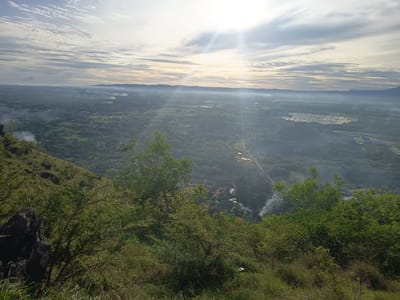 This image provides an elevated panoramic view of the landscape surrounding the city of Geundring, Indonesia, captured from a natural hillside. The sky is partially covered by scattered altocumulus and cirrus clouds, with prominent sun flares originating from the upper right quadrant, indicating bright light conditions. The immediate foreground is dominated by lush green vegetation, including tall grasses, shrubs, and trees, with a dark, rugged rock formation visible on the far left.
Below the vantage point, the midground reveals a vast expanse featuring a blend of urban development and agricultural land. Numerous buildings and residential areas are visible, interspersed with large patches of dense green foliage and cultivated fields, some appearing as water-filled terraced paddies. Several distinct plumes of white smoke are rising from different points within this developed area, suggesting localized burning activities. Roads and pathways can be observed winding through the terrain. The background consists of a hazy horizon line, behind which a range of distant mountains or hills is faintly visible. No individuals are discernible within the frame.
