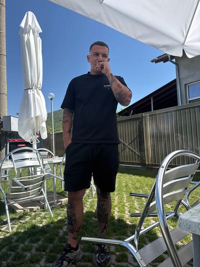 A man is standing outside in a yard with grass and stone pavers. He is wearing a black t-shirt, black shorts, and sneakers. He has tattoos on his arms and legs. There are metal chairs and tables around him, and a closed white umbrella is to his left. A fence is behind him, and a building with a roof is visible further back. The sky is blue. The location is described as the city of Kozarsko, Bulgaria.