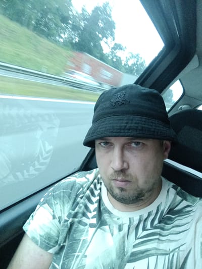 The image shows a man wearing a black bucket hat and a t-shirt with a palm leaf pattern, seated in what appears to be a vehicle. The man is looking directly at the camera with a serious expression. Through the window, a blurry green landscape with trees and a red structure is visible, suggesting movement, possibly on a road or a race track. The sky appears overcast, indicating a cloudy day. The interior of the vehicle is dimly lit, with part of a black seat visible in the background. The man has short brown hair and a light beard. There is no visible text in the image. The location context provided is Třemošnice, Czechia, which suggests the scene is likely taking place in that region. The overall impression is a candid, possibly casual, snapshot taken while traveling.