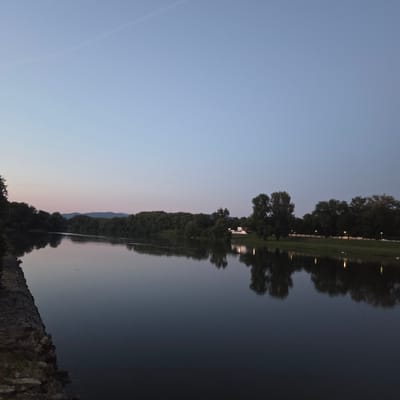 The image depicts the Váh River in Piešťany, Slovakia, during twilight. The sky is a pale blue transitioning to a soft pink hue near the horizon. On the left bank, a stone embankment is visible, leading into a dense line of trees that stretches along the river. On the opposite bank, a park-like setting features manicured grass, several trees, and a white building with illuminated windows, suggesting it might be a public facility or residence. Streetlights are visible along the edge of the park, casting reflections on the calm surface of the river. The water is dark and reflects the trees, lights, and the sky. In the distance, a faint outline of hills can be seen against the horizon. The scene is tranquil and captures the natural beauty of Piešťany at dusk.