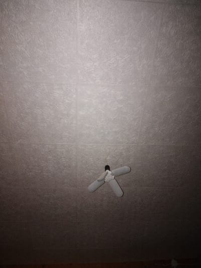 The image depicts a textured ceiling with a patterned surface, likely wallpaper or textured paint. A close-up view of the ceiling reveals a subtle, wavy texture. In the center of the frame, a white ceiling fan with three blades is suspended. The fan appears to be a modern design, possibly a LED ceiling fan light, with each blade shaped like a large, elongated petal. The lower portion of the image transitions to darkness, with a hint of wooden flooring visible at the very bottom edge. The scene is indoors, and the lighting is dim, casting shadows and creating a subdued atmosphere. There are no people or any specific landmarks that would indicate a location in Brish, Russia. The image focuses solely on the ceiling and the suspended fan.