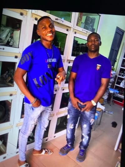 Two men pose in a commercial setting. Both wear blue shirts and jeans. The top man has dark jeans and blue sneakers, and he has a gold watch on his left wrist. The bottom man’s shirt has the word “London” on it, and he has lighter jeans with sandals. Behind them are shelving units, possibly displays of goods. The image is slightly rotated, and the lighting is bright.