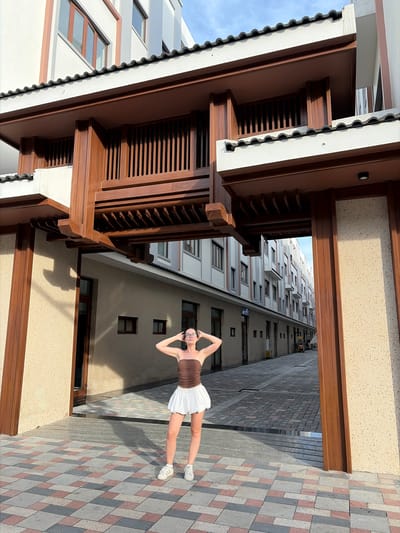 A young woman with dark hair and glasses stands in the middle of a paved walkway between two buildings. She is wearing a brown strapless top and a white skirt, and white sneakers. Her hands are behind her head. She is positioned under a wooden architectural structure with intricate latticework. The walkway is paved with a pattern of red, grey, and beige tiles. The buildings on either side have light-colored facades and multiple windows. The scene appears to be an outdoor area in a city.