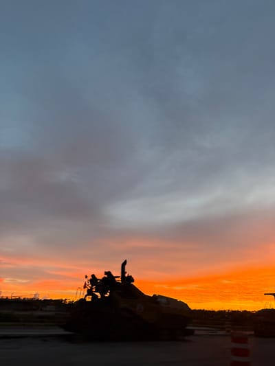 The image depicts a silhouette of construction equipment against a vibrant sunset sky. The sky displays a gradient of colors, transitioning from a pale blue at the top to fiery oranges and reds near the horizon. Wispy clouds are visible throughout the sky, illuminated by the setting sun. In the foreground, a large piece of construction machinery, possibly a bulldozer or excavator, is silhouetted in dark black. Its complex structure, including a bucket and engine components, is discernible against the bright sky. To the right, part of another piece of equipment and orange and white traffic cones are also visible, suggesting an ongoing construction site. The background shows a faint outline of trees and possibly some buildings, indicating an urban or suburban environment. The overall scene captures a moment of transition between day and night, with the industrial elements of the construction site juxtaposed against the natural beauty of the sunset. The location is the city of North Port, United States.