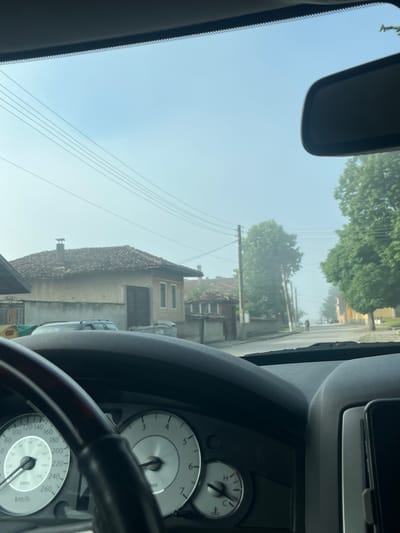 The image, taken from inside a vehicle, shows a view through the windshield of a street in Ivanski, Bulgaria. The scene is hazy, suggesting either early morning fog or light rain. A traditional Bulgarian house with a tiled roof and a chimney is visible on the left. The street is lined with trees and utility poles, with power lines crisscrossing overhead. A few cars are parked along the road, and a single person is walking in the distance. The dashboard of the car, including the steering wheel and speedometer, is in the foreground.