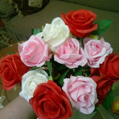 The image shows a close-up, overhead view of a bouquet of artificial roses. The bouquet contains a mix of red, pink, and white roses, with green artificial leaves interspersed. The roses are arranged in a dense cluster. In the foreground, a portion of a person's arm and hand is visible, suggesting the bouquet is being held or presented. The background is out of focus and appears to be an indoor setting with furniture. The lighting is somewhat dim, creating a muted atmosphere. There is no visible text in the image. The context of Ashtarak, Armenia, is provided as location information.