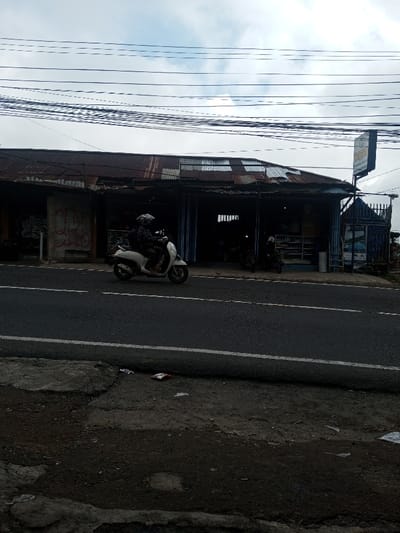 A moped is traveling on a road in front of a rustic building with a corrugated iron roof. Power lines crisscross overhead. The building appears to be a small shop or market. There is trash strewn on the shoulder of the road. The sky is overcast and the overall scene is somewhat gloomy.