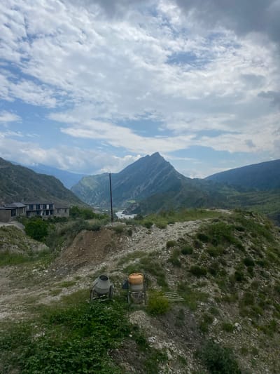 The image shows a mountainous landscape with a cloudy sky. In the foreground, there are two concrete mixers on a grassy, rocky slope. Behind the concrete mixers, there is a dirt path and some vegetation. Further in the background, there are several buildings, a river, and more mountains. The scene appears to be in or near the village of Chalda, Russia, based on the architectural style of the buildings and the mountainous terrain. The lighting suggests it is daytime, with the sun partially obscured by clouds.