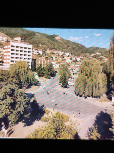 The image depicts a high-angle view of an intersection in the city of Devin, Bulgaria. A large, white multi-story building stands prominently in the background. The landscape includes a mix of buildings, trees, and a mountain in the distance. Several people are visible crossing the street at the intersection. Cars are parked near the trees.