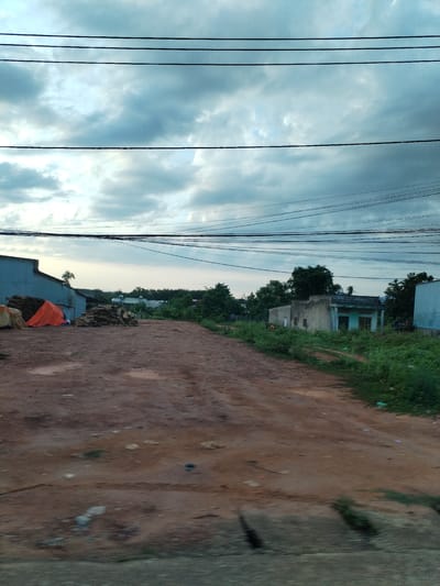 The image depicts a rural scene in Đăk Xú, Vietnam. An unpaved, reddish-brown dirt road stretches into the distance. Overhanging electrical wires cross the sky, which is overcast with grey clouds. To the left, there is a corrugated metal building with an orange tarp partially covering a pile of wood. Another pile of wood is next to it. The side of the road has green plants. The building has a window, and a door to the right of the window.
