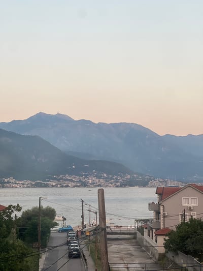 The image captures a scenic view of the city of Bijela, Montenegro, looking out towards the bay and mountains. In the foreground, a street slopes down towards the water, lined with parked cars and a residential building on the right. The building is a light gray, two-story structure with a red-tiled roof, featuring multiple balconies and windows. Further down the street, a body of water is visible, with several small boats docked near the shore. Beyond the immediate foreground, a town stretches along the coastline, with buildings clustered together. In the background, majestic mountains rise, their peaks softened by a hazy atmosphere. The sky above transitions from a pale blue at the top to a soft, peachy hue near the horizon, suggesting it might be dusk or dawn. The overall scene conveys a peaceful coastal town atmosphere.