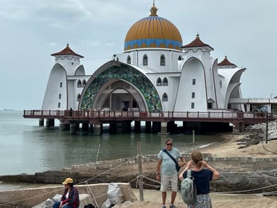 The Malacca Straits Mosque, a white building with a blue and gold dome, stands over the water on stilts. A large arched entrance features intricate stained glass. In the foreground, a low stone wall runs along the beach. Three individuals occupy the foreground: one seated on the wall, one standing with hands near their head, and another nearby with a shoulder bag. The sky is overcast, and a ship is visible in the distance.