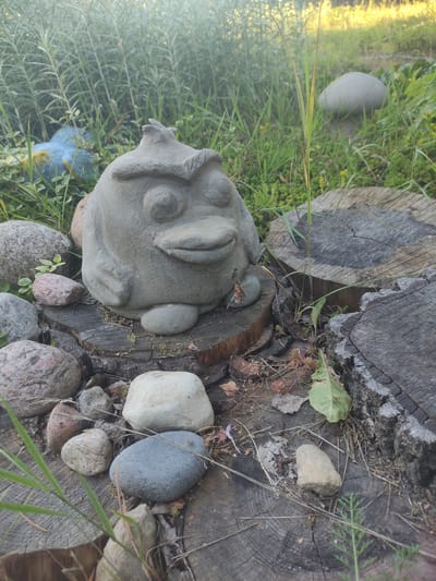 A concrete garden statue resembling a stylized bird or creature sits on a tree stump in an outdoor setting. The statue has a rounded body, large circular eyes, and a wide mouth. It is surrounded by various stones, a few pieces of wood, and lush green vegetation. In the background, more greenery and what appears to be another concrete garden ornament, possibly a mushroom, are visible. The overall scene suggests a garden or yard decoration in the Верамейскі сельскі Савет, Belarus.