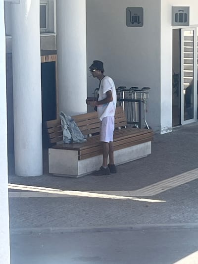 A man is standing next to a wooden bench with a jacket draped over it, smoking a cigarette. He is wearing a black bucket hat, a white t-shirt, and light pink shorts. He has on dark shoes. Behind him, luggage carts are lined up. The background shows a building with large white pillars, a glass door, and signage indicating luggage carts and luggage. This appears to be an outdoor area, possibly an airport or train station. The scene is brightly lit by sunlight, casting shadows on the ground. The image does not provide any specific clues to place it in Viseu, Portugal.