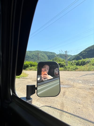 A reflection in a side-view mirror shows a young woman holding a phone in her right hand, taking a selfie. She is wearing a white top and has dark hair. Next to her, a white dog with its mouth open and tongue out is visible. The background in the mirror is a green, tree-covered mountainside with a clear blue sky above. Power lines and a utility pole are visible in the distance. The vehicle appears to be on a gravel road. The image is taken from inside the vehicle, looking out towards the side mirror. The approximate location is Krasnoye, Russia.