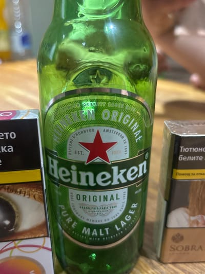 The image shows a close-up of a green glass bottle of Heineken beer. The bottle has a label that reads "Heineken Original" in large letters, with "Premium Quality" and "Amsterdam" also visible. The label also features a red star. The bottle is on a wooden surface, likely a table. To the left of the bottle, a portion of a cigarette pack is visible, with text that appears to be in Cyrillic script. To the right of the bottle, another cigarette pack is visible, also with Cyrillic text, and an image of a person. The background is blurred, but appears to be an indoor setting with some light sources. The image is a still photograph and does not depict any actions or interactions. There is no information in the image to suggest the location is Boston, United Kingdom.