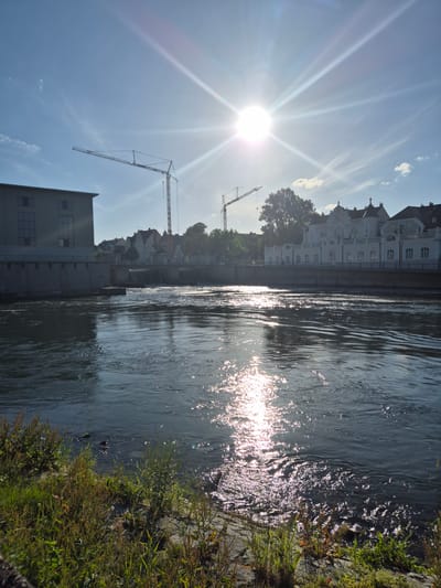 This image shows a river in Landshut, Germany, with buildings and construction cranes in the background. The sun is shining brightly, creating a glare on the water. The riverbank is covered in grass and other vegetation. In the distance, there are several buildings, including a large white building with ornate architecture. Two construction cranes are visible in the sky, suggesting that there is ongoing construction in the area. The water in the river is flowing, and there are ripples and reflections on its surface. The overall scene is one of a sunny day in a city with both historical and modern elements.