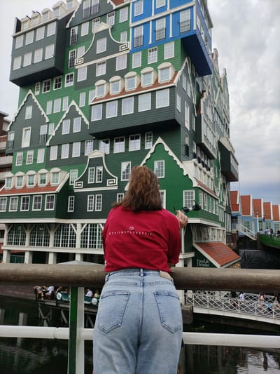 A person, viewed from behind, stands on a wooden walkway, looking towards a uniquely designed, multi-story building. The building is a striking assembly of green, blue, and white sections, each resembling traditional Dutch houses stacked and cantilevered on top of each other. Many windows are visible on the facade. The person is wearing a red long-sleeved shirt with white text and light blue denim jeans. The text on the shirt appears to read "DTSIINCTLVESTYLE.".
Below the building, a body of water can be seen, reflecting the structures. In the distance, a bridge crosses the water, with several people walking across it. Other smaller buildings with red-tiled roofs are also visible along the water's edge, adding to the picturesque scene. The sky is overcast, suggesting a cloudy day. The architecture strongly suggests a location in the Netherlands, specifically Zaandam, known for its distinctive wooden house architecture. The overall mood is one of appreciation for the unusual and colorful urban landscape.