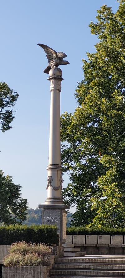 This is a vertical, eye-level, medium shot of a monument in Wattens, Austria. The monument is a tall, silver column topped with a statue of an eagle with its wings spread. The column has decorative elements, including two lion heads on either side near the base of the column. At the base of the monument, there is a granite plaque that reads "MAGNARES LIBERTAS" in white lettering. The monument is surrounded by green bushes and trees, with a clear blue sky in the background. The monument appears to be located in a park or public space, as evidenced by the stone seating area and steps in the foreground. The lighting suggests it is daytime.