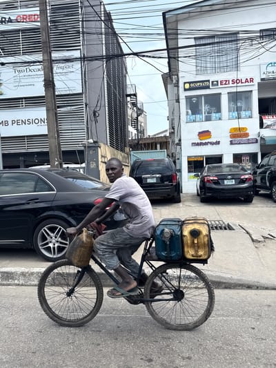 The image depicts a man on a bicycle in what appears to be Falomo, Nigeria, based on the signage visible in the background. The man is transferring liquid, presumably fuel, from a yellow jerrycan into a black car. His bicycle has two jerrycans strapped to the back. The setting is a street, with buildings displaying advertisements for businesses such as "EZI SOLAR," "MB PENSIONS," and "GCUPS LTD.". Several cars are parked along the street. Power lines are also prominently featured.