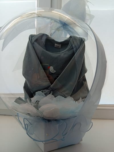 A baby onesie is displayed inside a clear, round balloon that is part of a gift presentation. The onesie is gray and has a small white bird with an orange beak printed on the front. A size tag that reads "6-9M" is visible on the inside collar. The balloon is supported by a light blue gift box adorned with decorative netting and blue cord. The overall scene appears to be a gift for a baby, possibly a newborn. The background is out of focus, showing a white surface that could be a window blind or curtain. The image is taken from a slightly low angle, looking up at the gift. There are no people visible in the image. The context of Tomai, Moldova, is not evident in the visual information provided.