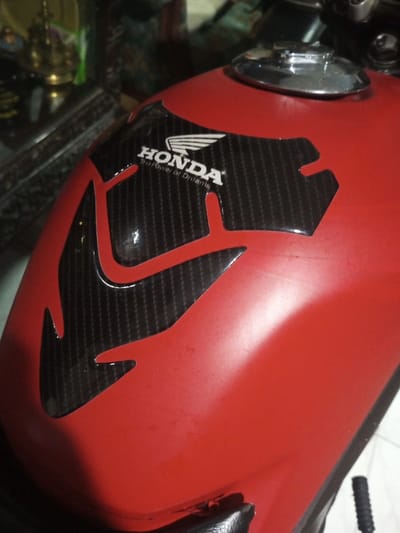 A close-up photograph displays the red fuel tank of a motorcycle, located indoors in B. Nam Ngao, Laos. The tank exhibits a matte finish with minor surface abrasions. A black, glossy tank pad, featuring a carbon-fiber weave pattern and an angular design, is adhered to the upper-front surface of the tank. Centered on this tank pad is the white "HONDA" logo with a winged emblem, above the white text "The Power of Dreams." A chrome-plated fuel cap is positioned on the top-right aspect of the tank. The background is out of focus, showing a partial brass-colored decorative object and indistinct dark furnishings to the left, with light-colored floor tiles visible in the lower right. No individuals are present in the image.