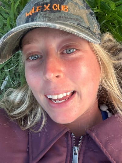 A person with fair skin, blonde hair, and blue eyes is lying on their back in a grassy area. They are wearing a camouflage baseball cap with the words "WOLF X CLUB" embroidered in orange. They are also wearing a maroon or purple jacket with a visible zipper. The location is impossible to determine based on the image.