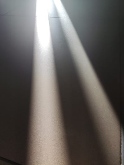 The image displays a close-up, overhead view of a floor surface, likely indoors, characterized by distinct light and shadow patterns. Two parallel beams of bright sunlight cut diagonally across the frame from the upper left corner, illuminating a textured, granular surface that appears to be tiled or concrete. The light is intense, creating a gradient from bright white in the illuminated areas to deep shadow on the right side of the image. Subtle grid lines are visible on the floor, suggesting a tiled surface, and faint marks or specks are scattered across it. The scene is devoid of any human presence or identifiable objects other than the floor itself and the light. There is no information in the image that would allow for the determination of its location within the city of Polatsk, Belarus.