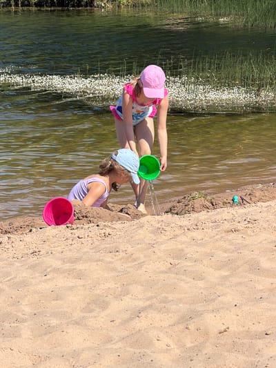 Two young girls are playing at the edge of a body of water on a sunny day. The girl on the left, wearing a pink hat and a multi-colored swimsuit, is sitting in the water and holding a green bucket. She is pouring water from the bucket. The girl on the right, wearing a light purple swimsuit and a blue baseball cap, is sitting on the sandy shore and playing with sand. She has a pink bucket next to her. The water appears shallow and calm, with green vegetation growing along the bank. The sand is light brown and rippled from the water. The scene suggests a leisurely day at the beach or lake, likely during the summer. The lighting indicates it is daytime. No text is visible in the image. The location context of Vētras, Latvia, places this scene in a natural outdoor environment.