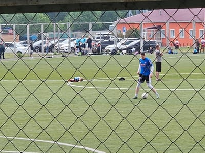 The image depicts an outdoor football match on a synthetic turf field. Several individuals are present on the field, some actively participating in the game and others observing. In the foreground, a player in a blue jersey is kicking a soccer ball. To the left of this player, another individual in a blue and red uniform is lying on the ground. Other players and spectators are visible in the background, some near parked cars and a red building with a light-colored roof, possibly a sports facility. The scene appears to be taking place in the city of Sosnovoborsk, Russia, given the visual context of the surrounding environment, which suggests a typical urban or semi-urban setting. The overall atmosphere seems to be that of a casual or amateur football game.