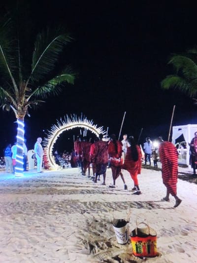 This image depicts a nighttime outdoor event on a sandy area resembling a beach, flanked by palm trees. The environment is inconsistent with the inland city of Dodoma, Tanzania, suggesting a coastal or beach-themed setting.
In the foreground, two decorative buckets rest on the sand; one is white with black text, and the other is a colorful, patterned basket-like container. The ground is light-colored sand with visible footprints and undulations.
In the mid-ground, a procession of approximately eight individuals, predominantly male, walks from right to left in a line. They are dressed in red, patterned shukas or cloths reminiscent of Maasai attire, and most carry slender staffs. They appear to be approaching or passing through a large, circular archway illuminated with numerous small white lights, creating a spiky, radiant effect.
To the far left, a palm tree is wrapped with vibrant blue string lights. Several individuals in contemporary clothing are standing near this tree, observing the scene. One man in a light-colored shirt is prominent. Further back, another group of people is visible near the archway and beyond it.
To the far right, part of a white tent or canopy structure is visible. The sky is dark, indicating night. Artificial lighting illuminates the immediate area and the main subjects.