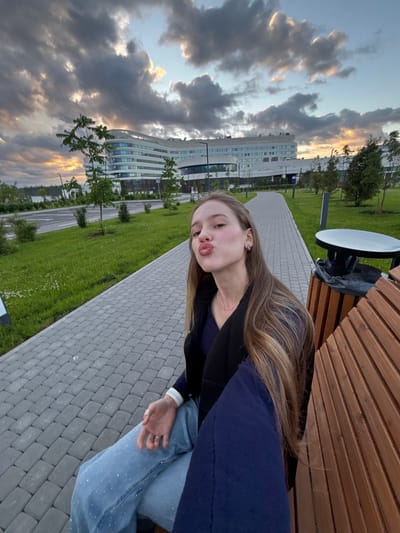A young woman with long, blonde hair is sitting on a wooden bench, pouting her lips. She is wearing a dark jacket over a dark blue top and light blue jeans adorned with small silver rhinestones. Her left hand rests on her lap, and a ring is visible on her finger. Behind her, a paved walkway lined with grass and small trees leads towards a modern, white, curved building with multiple windows. The sky above is filled with dramatic clouds, with hints of sunset colors peeking through. The location is the city of Solnechnoye, Russia.