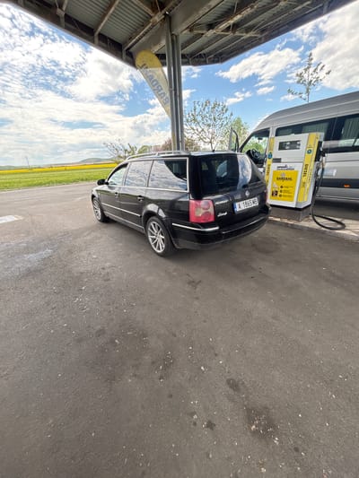 The image shows a black Volkswagen Passat Variant parked at a gas station in Sadievo, Bulgaria. The license plate of the car reads "A 1865 MB." The car is parked next to a white van and a yellow diesel pump labeled "DIESEL + BDC," with the Bardahl logo visible. The pump is under a canopy. In the background, a field of yellow rapeseed flowers can be seen, with a cloudy sky above.